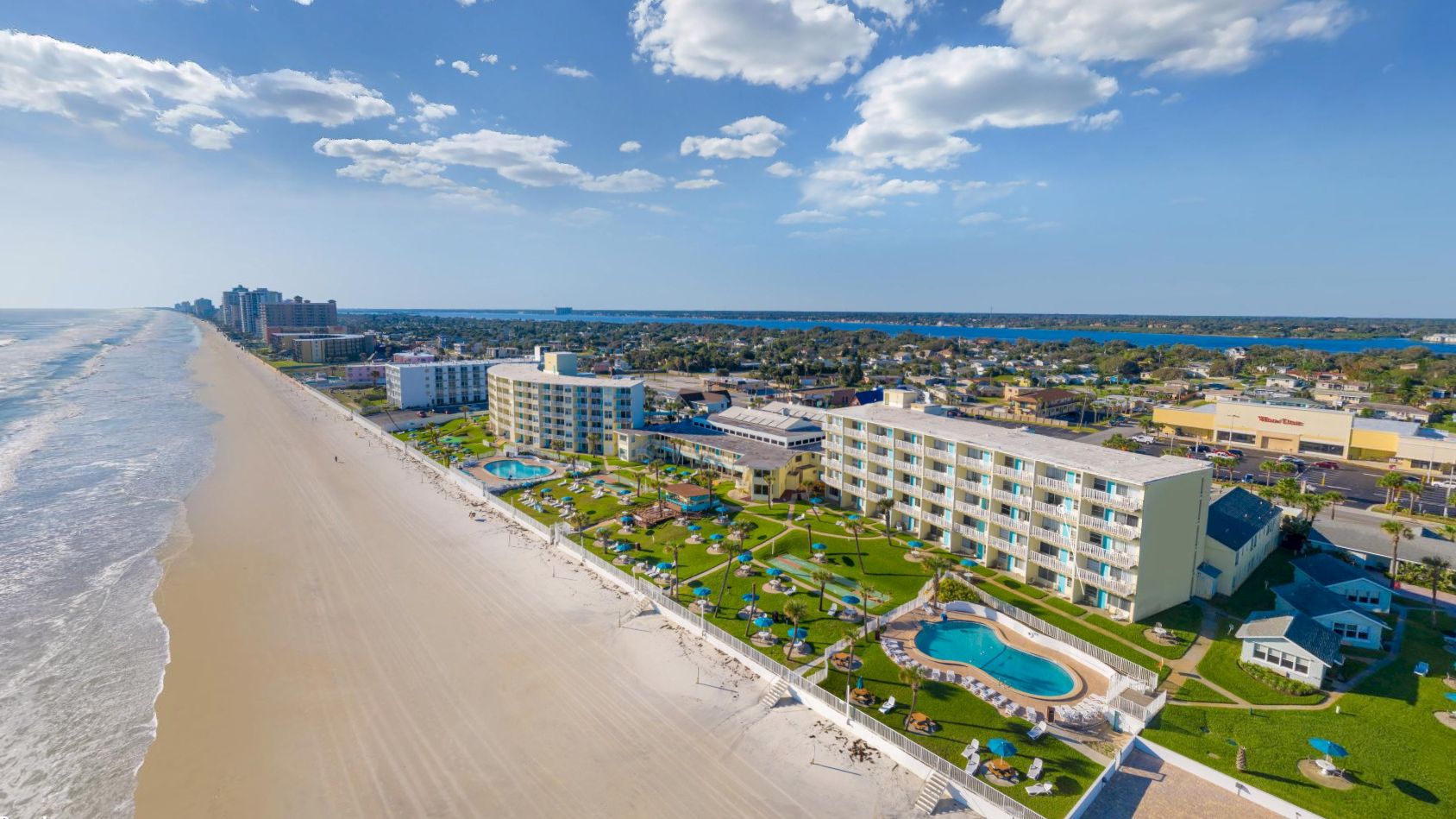 The image shows a coastal resort with multiple buildings, a swimming pool, and a beach extending along the shoreline under a partly cloudy sky.
