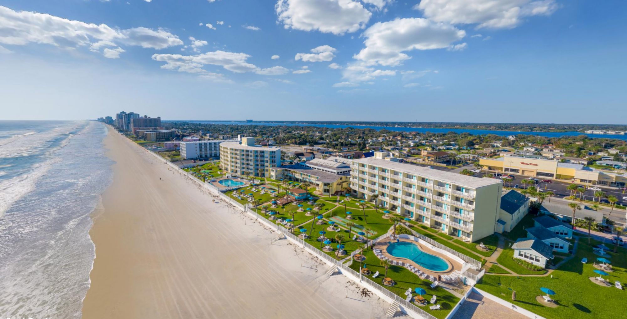 The image shows a coastal resort with multiple buildings, a swimming pool, and a beach extending along the shoreline under a partly cloudy sky.