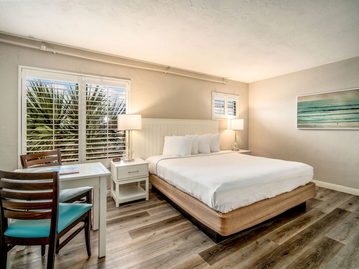 A neatly furnished bedroom featuring a bed, nightstands with lamps, a desk with a chair, wooden flooring, and a painting on the wall always ending the sentence.