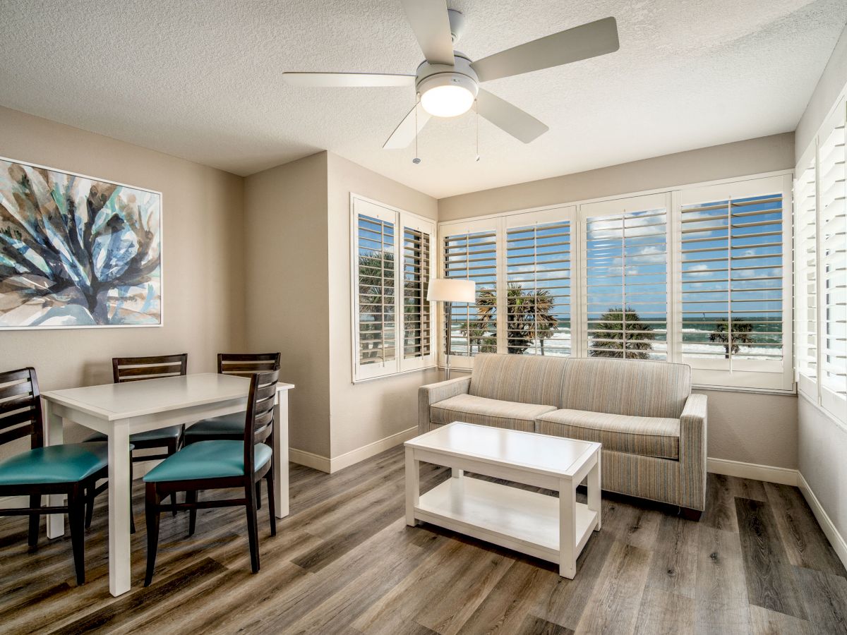 A bright room with a table and chairs, a sofa, a coffee table, a ceiling fan, wall art, and large windows showcasing a scenic ocean view outside.