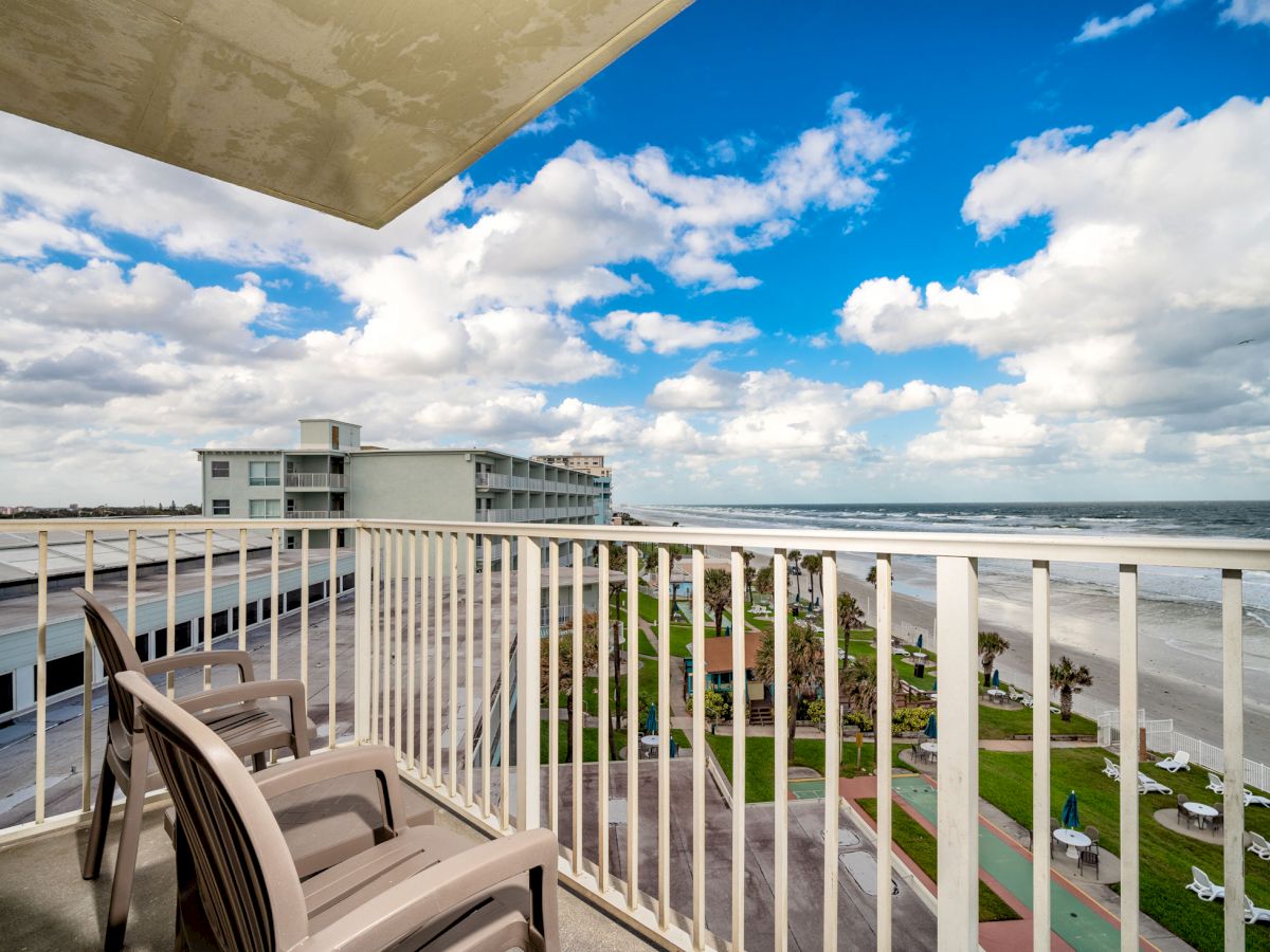 A balcony with two chairs overlooks a beachfront with a view of the ocean, a building, green spaces, and a partly cloudy sky in the background.