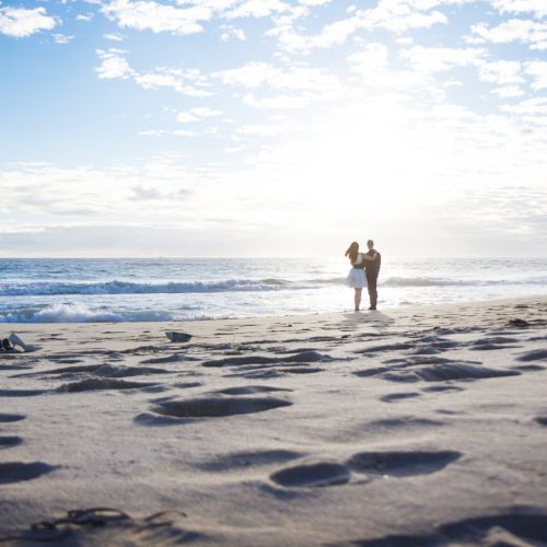 A couple is standing on a sandy beach near the ocean under a bright, cloudy sky with footprints in the foreground and waves in the background.