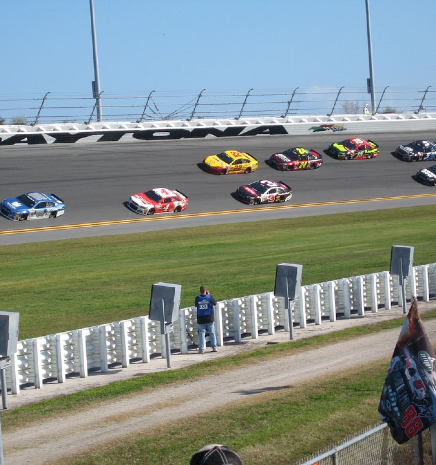 Race cars are on a track, with spectators and banners in the foreground, at Daytona International Speedway.