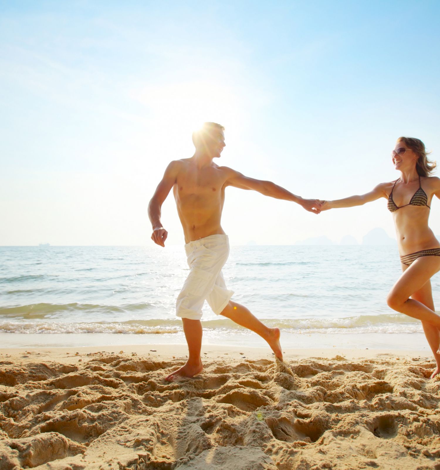 A couple in swimwear holds hands and runs along a sunny beach near the ocean, with a clear blue sky in the background.