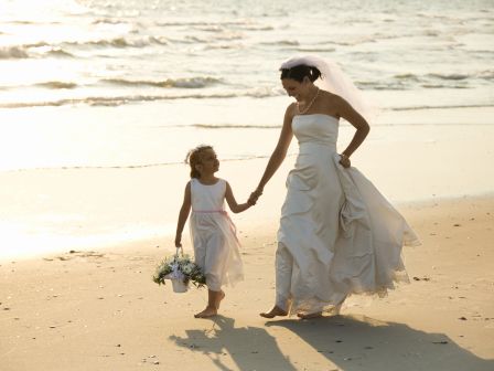 A bride and a young girl, likely a flower girl, dressed in white, walk hand in hand along a sandy beach by the ocean during sunset.