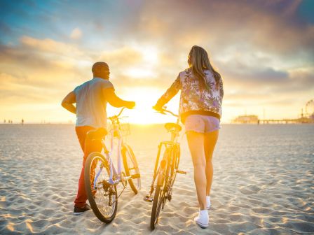 Two people walk their bicycles on a sandy beach during a picturesque sunset, with a ferris wheel visible in the background.