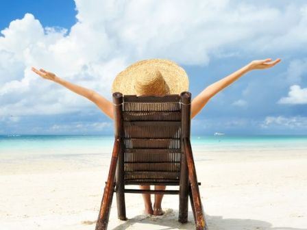 A person sits on a beach chair facing the ocean with their arms outstretched, under a cloudy sky.